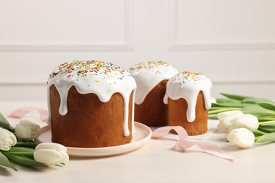Delicious Easter cakes with sprinkles and beautiful tulips on white wooden table indoors