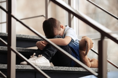 Sad little boy with toy sitting on stairs indoors