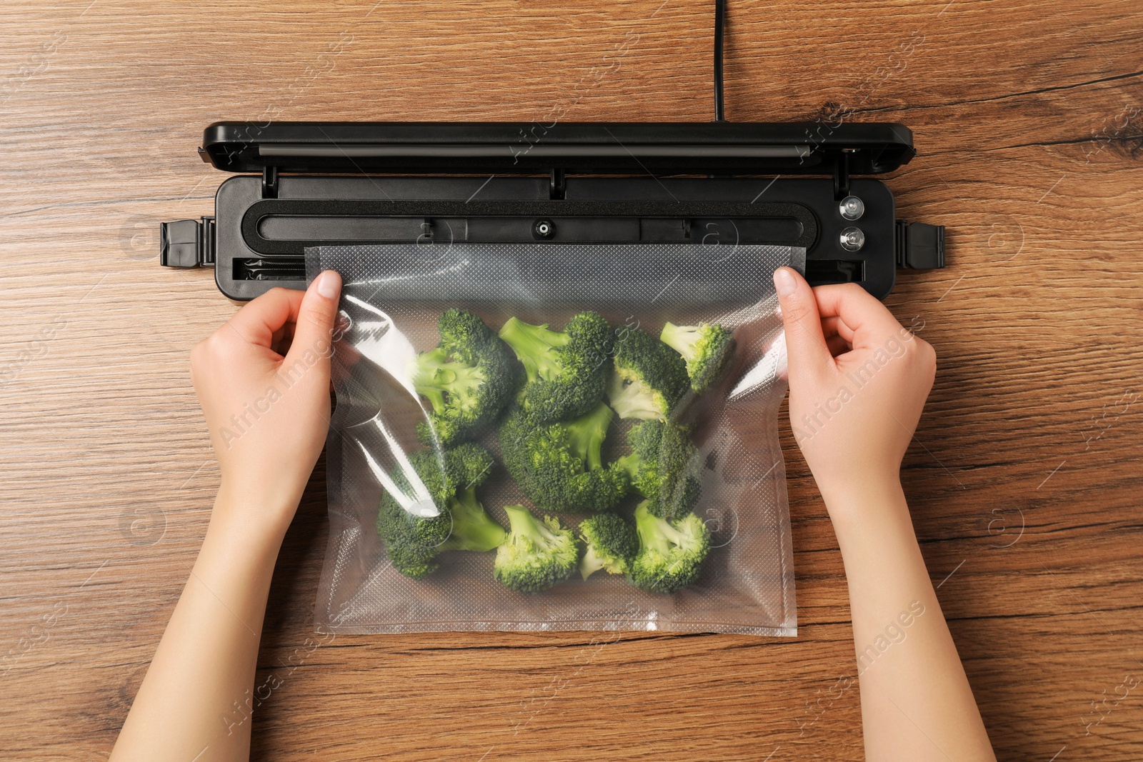 Photo of Woman using vacuum sealer at wooden table, top view. Green broccoli in pack