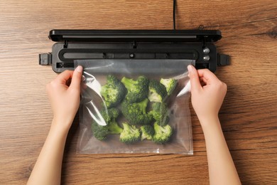Photo of Woman using vacuum sealer at wooden table, top view. Green broccoli in pack