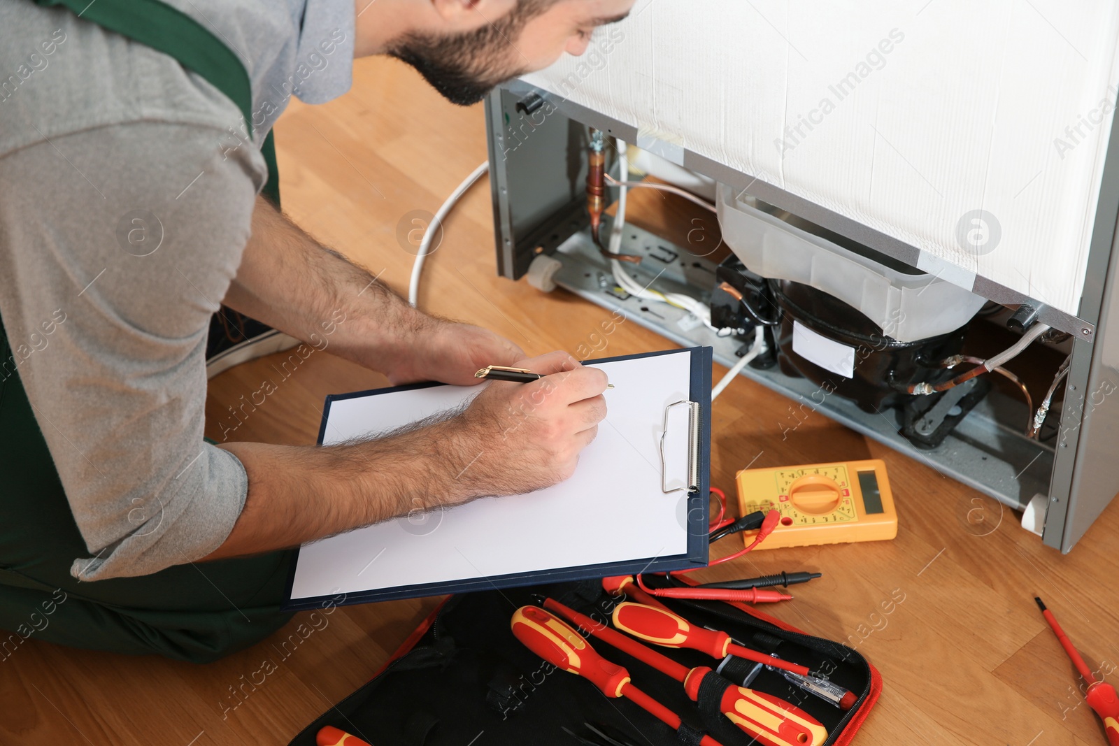 Photo of Male technician with clipboard examining broken refrigerator indoors, closeup