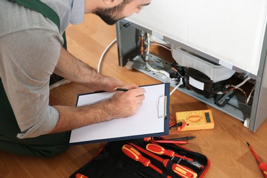 Male technician with clipboard examining broken refrigerator indoors, closeup