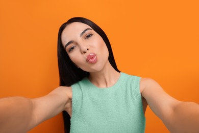 Beautiful young woman taking selfie while blowing kiss on orange background