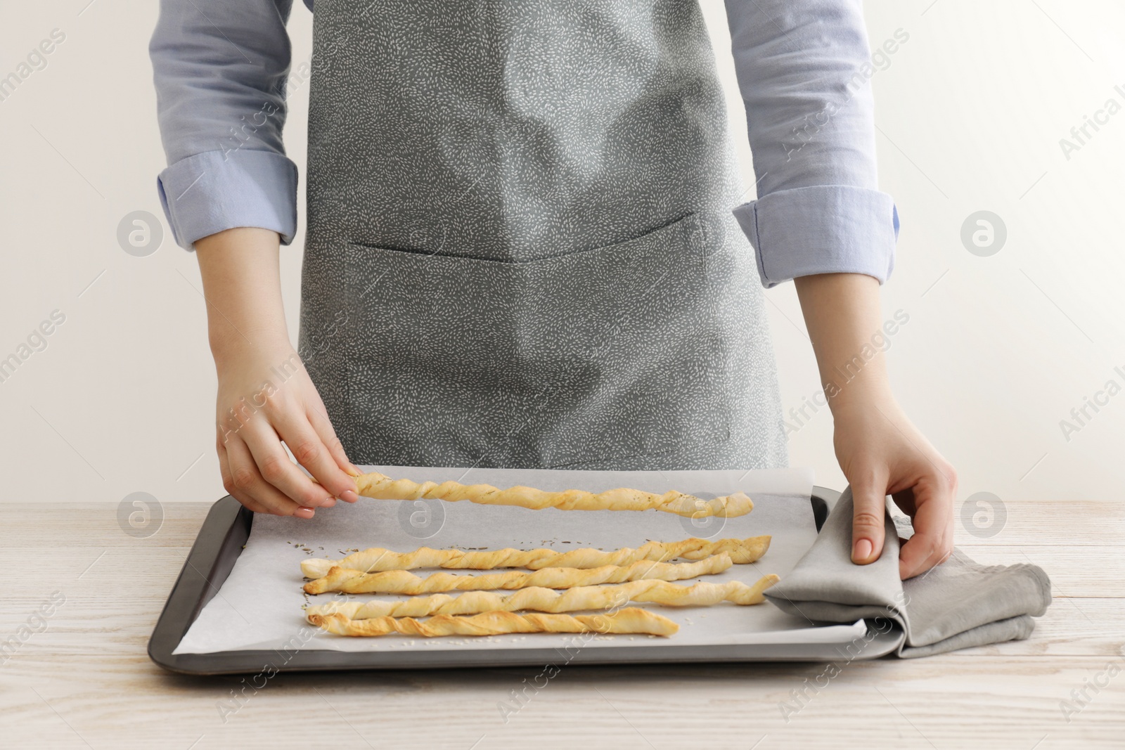 Photo of Woman near baking sheet with homemade breadsticks at white wooden table, closeup. Cooking traditional grissini