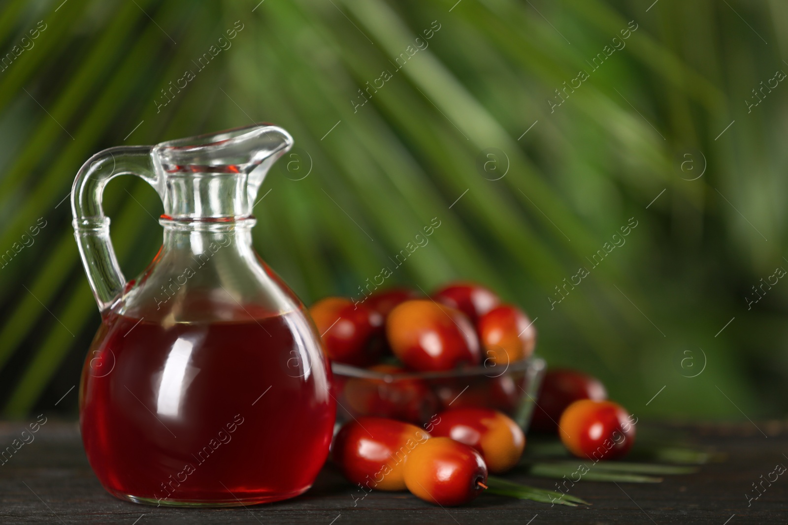 Photo of Palm oil in glass jug, tropical leaf and fruits on wooden table. Space for text