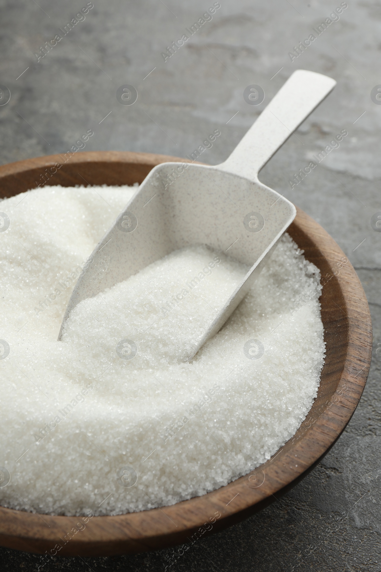 Photo of Granulated sugar in bowl and scoop on grey textured table, closeup