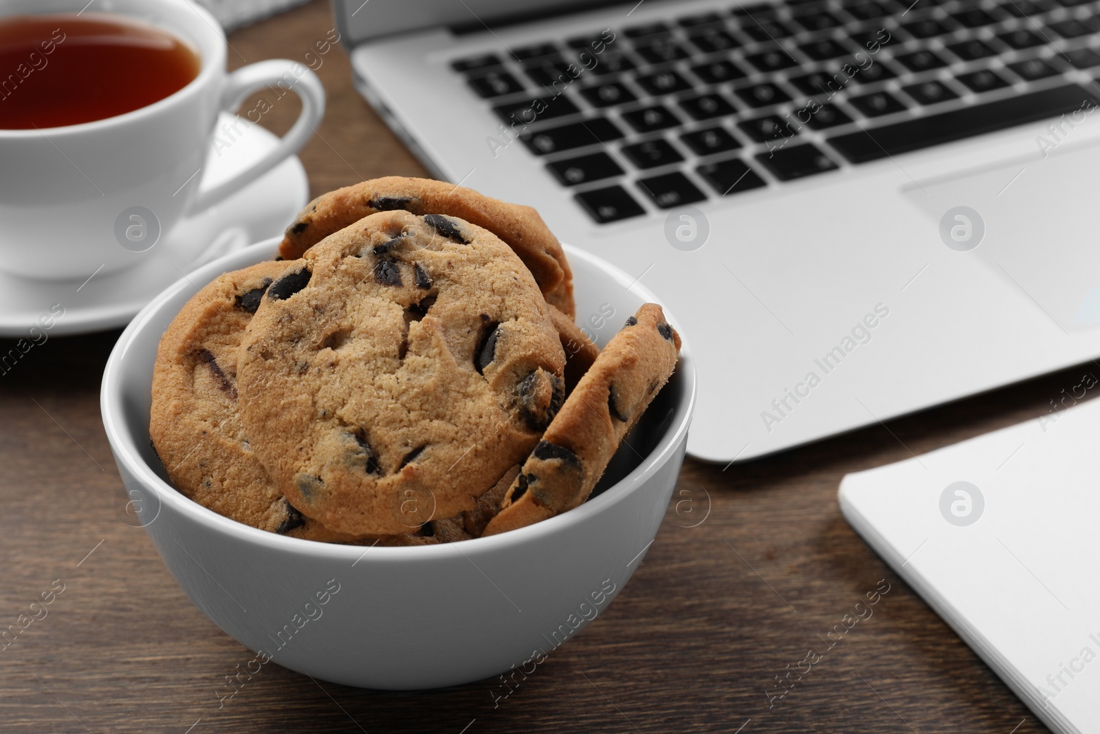 Photo of Chocolate chip cookies, cup of tea and laptop on wooden table, closeup. Space for text