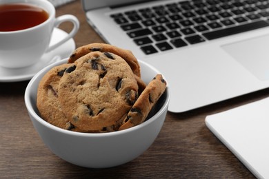 Photo of Chocolate chip cookies, cup of tea and laptop on wooden table, closeup. Space for text