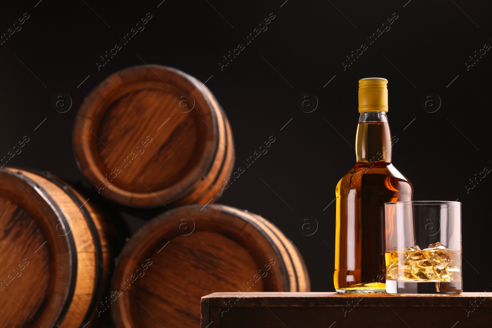 Photo of Whiskey with ice cubes in glass and bottle on wooden table near barrels against black background, space for text