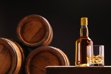 Whiskey with ice cubes in glass and bottle on wooden table near barrels against black background, space for text