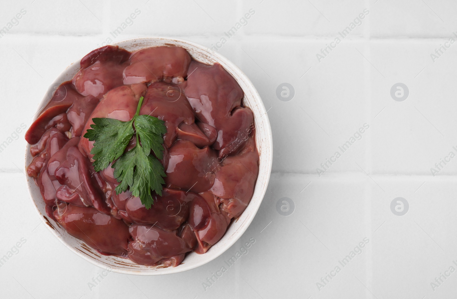 Photo of Bowl of raw chicken liver with parsley on white tiled table, top view. Space for text