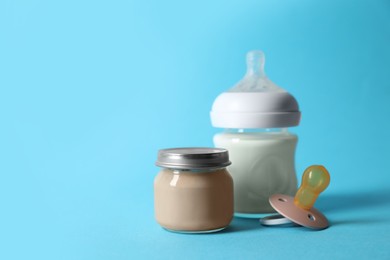 Photo of Jar with healthy baby food, bottle of milk and pacifier on light blue background. Space for text