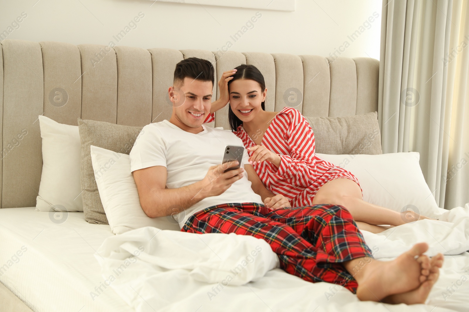 Photo of Happy couple in pyjamas using smartphone on bed at home