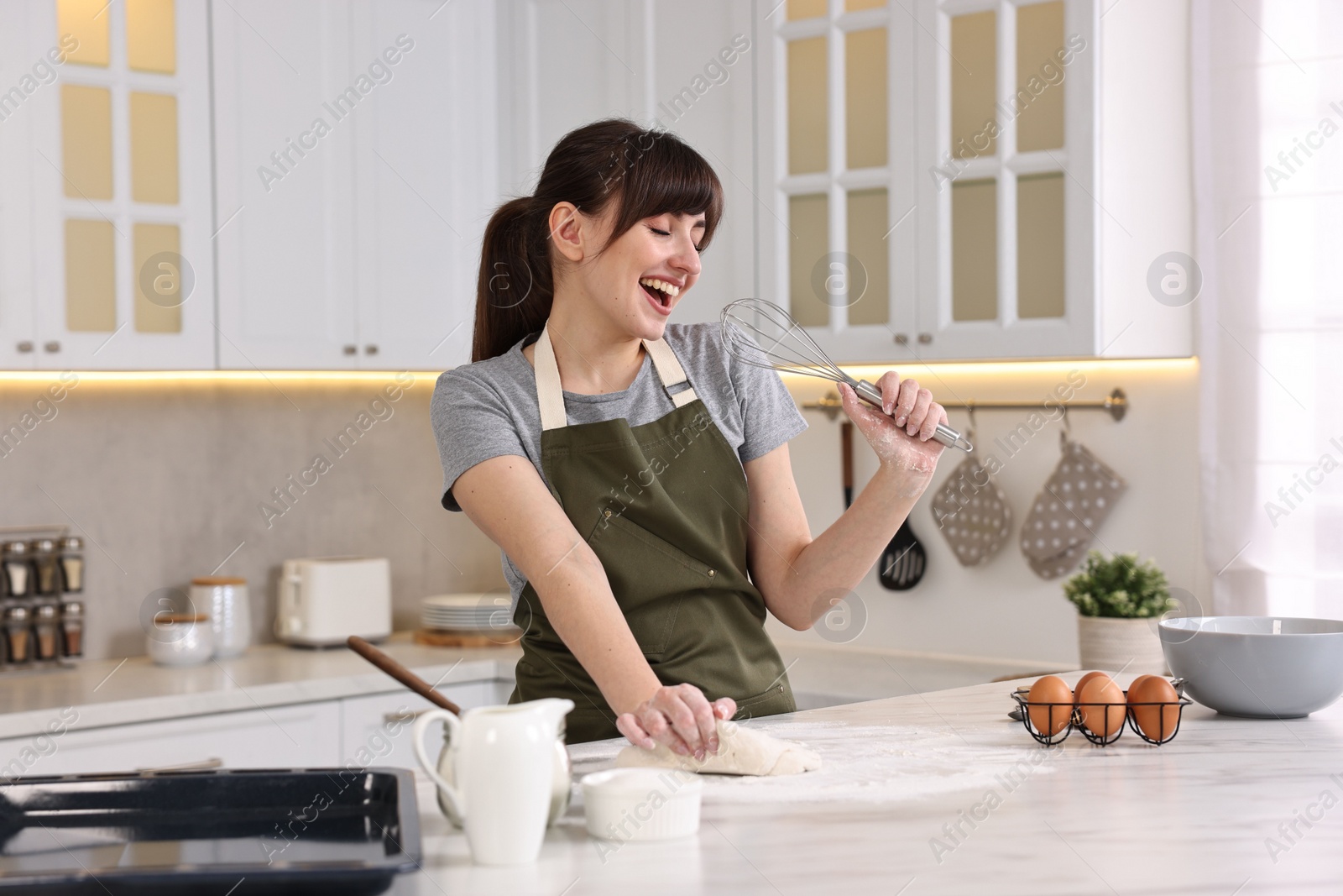 Photo of Happy young housewife with whisk having fun while cooking at white marble table in kitchen