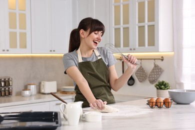 Happy young housewife with whisk having fun while cooking at white marble table in kitchen