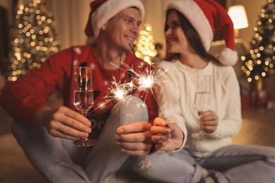 Photo of Couple in Santa hats holding sparkles and champagne glasses, focus on fireworks. Christmas celebration