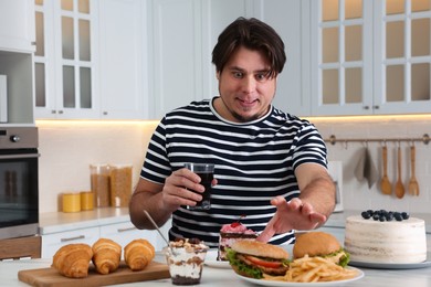 Photo of Hungry overweight man with glass of cold drink  taking burger from plate at table in kitchen