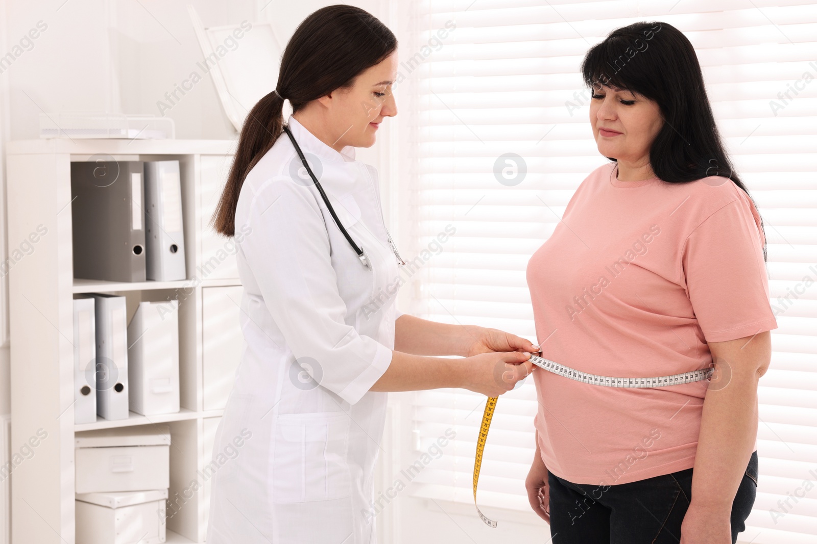 Photo of Nutritionist measuring overweight woman's waist with tape in clinic