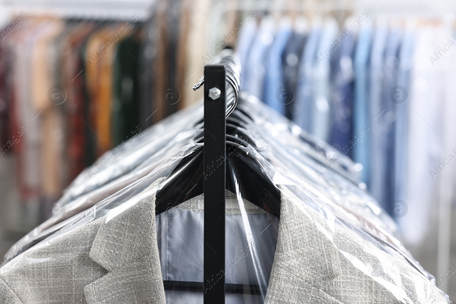 Photo of Dry-cleaning service. Many different clothes in plastic bags hanging on rack indoors, closeup