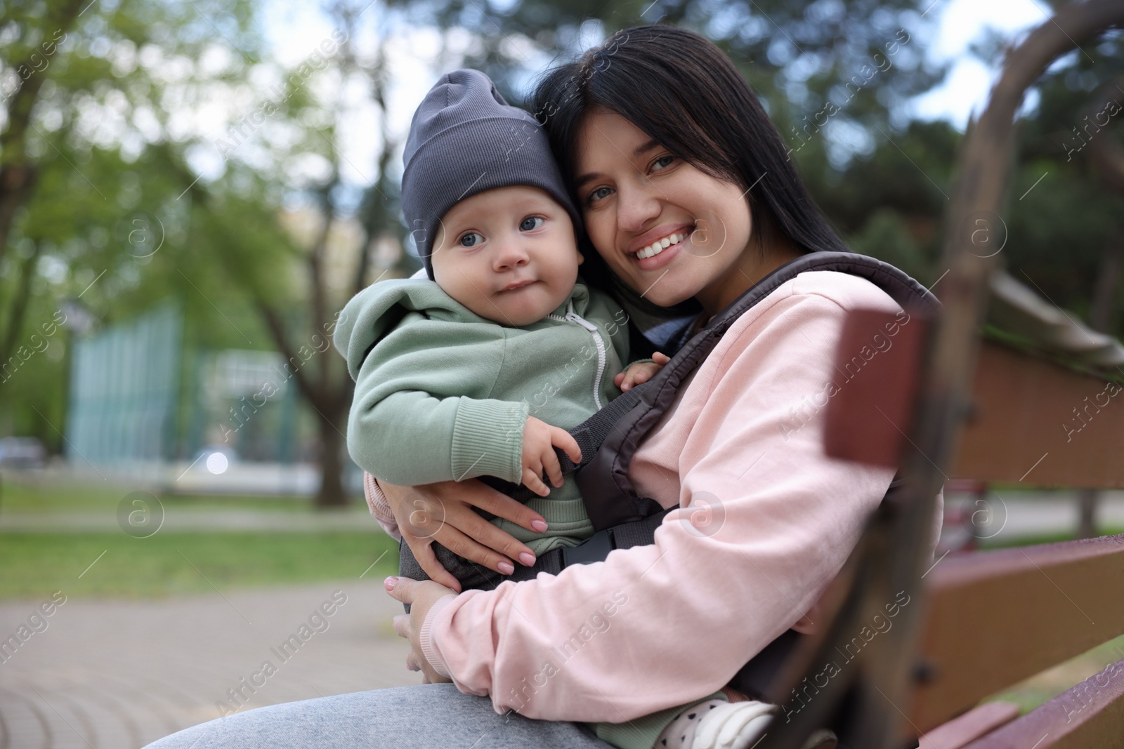 Photo of Mother holding her child in sling (baby carrier) on bench in park