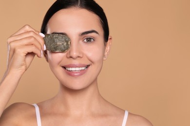 Photo of Beautiful young woman putting green tea bag on eye against beige background, space for text