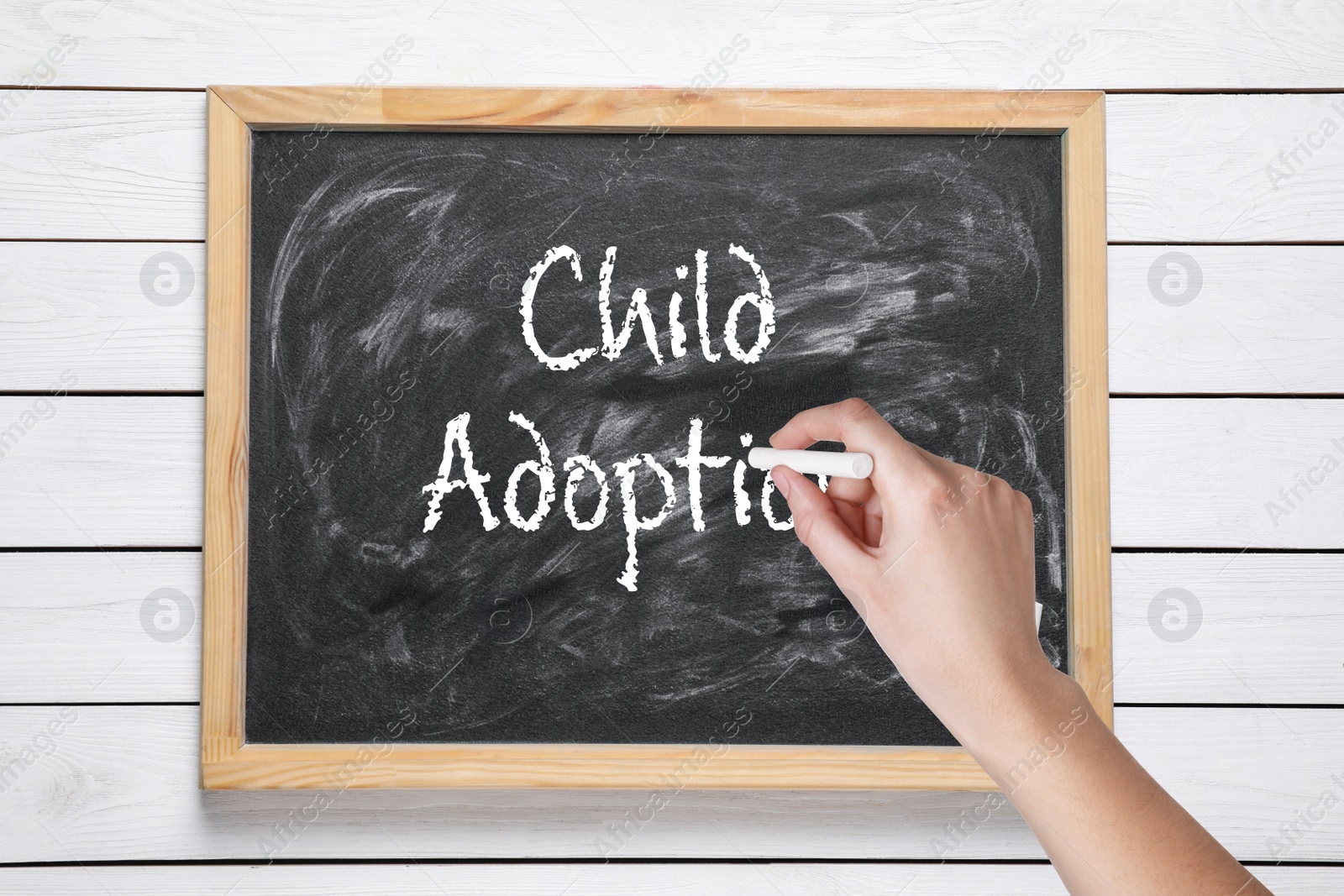 Image of Woman writing phrase CHILD ADOPTION on blackboard against white wooden background, closeup