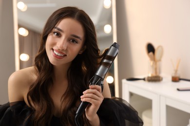 Photo of Smiling woman using curling hair iron at home. Space for text