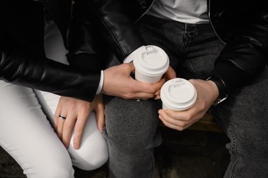 Lovely couple with coffee holding hands together while sitting on bench outdoors, closeup. Romantic date