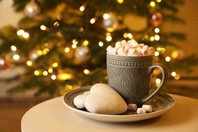 Photo of Cup of tasty cocoa with marshmallows and cookies on white table against blurred Christmas tree