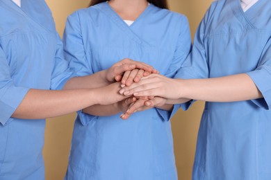 Photo of Nurses in medical uniforms putting hands together on light brown background, closeup