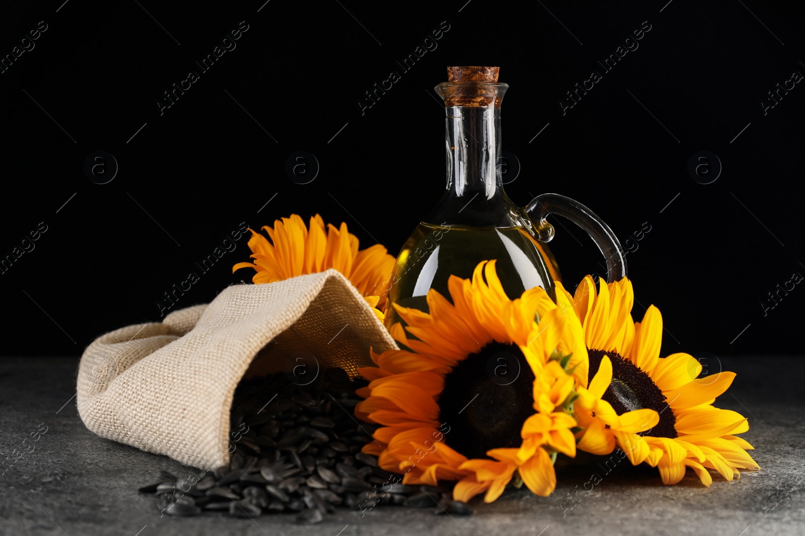 Photo of Sunflower cooking oil, seeds and beautiful flowers on grey table against black background