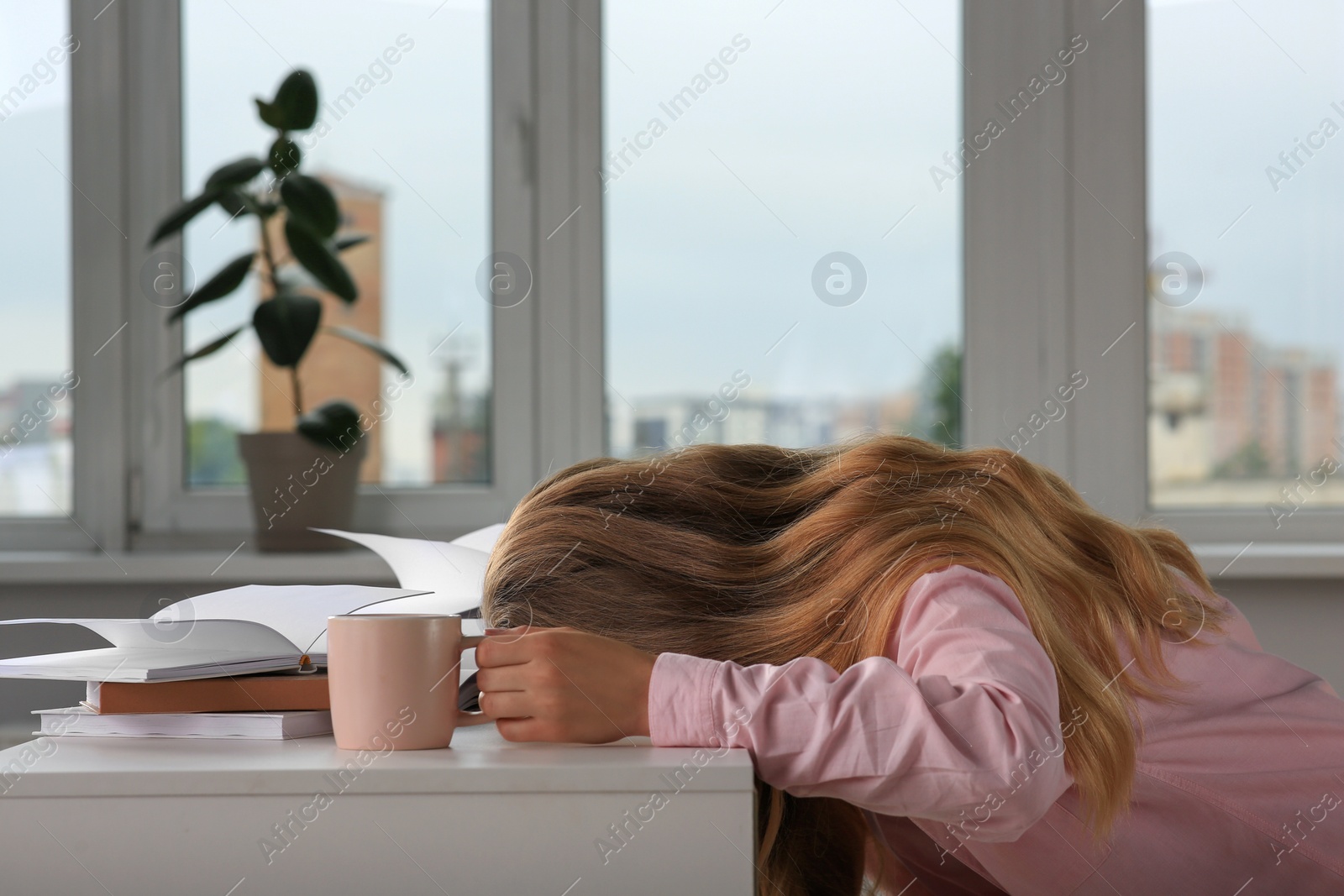 Photo of Young tired woman sleeping near books at white table in room