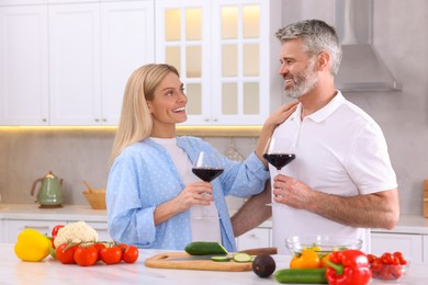 Photo of Happy affectionate couple with glasses of wine cooking together at white table in kitchen