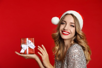 Happy young woman wearing Santa hat with Christmas gift on red background