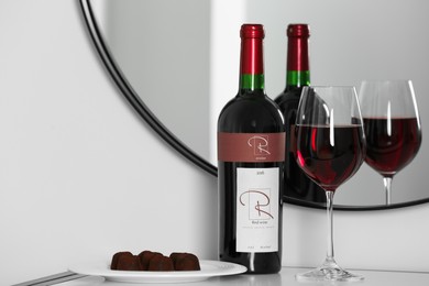 Photo of Bottle and glass of red wine with chocolate candies on table near mirror