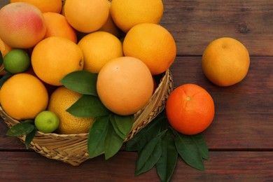 Wicker basket with different citrus fruits and leaves on wooden table, closeup