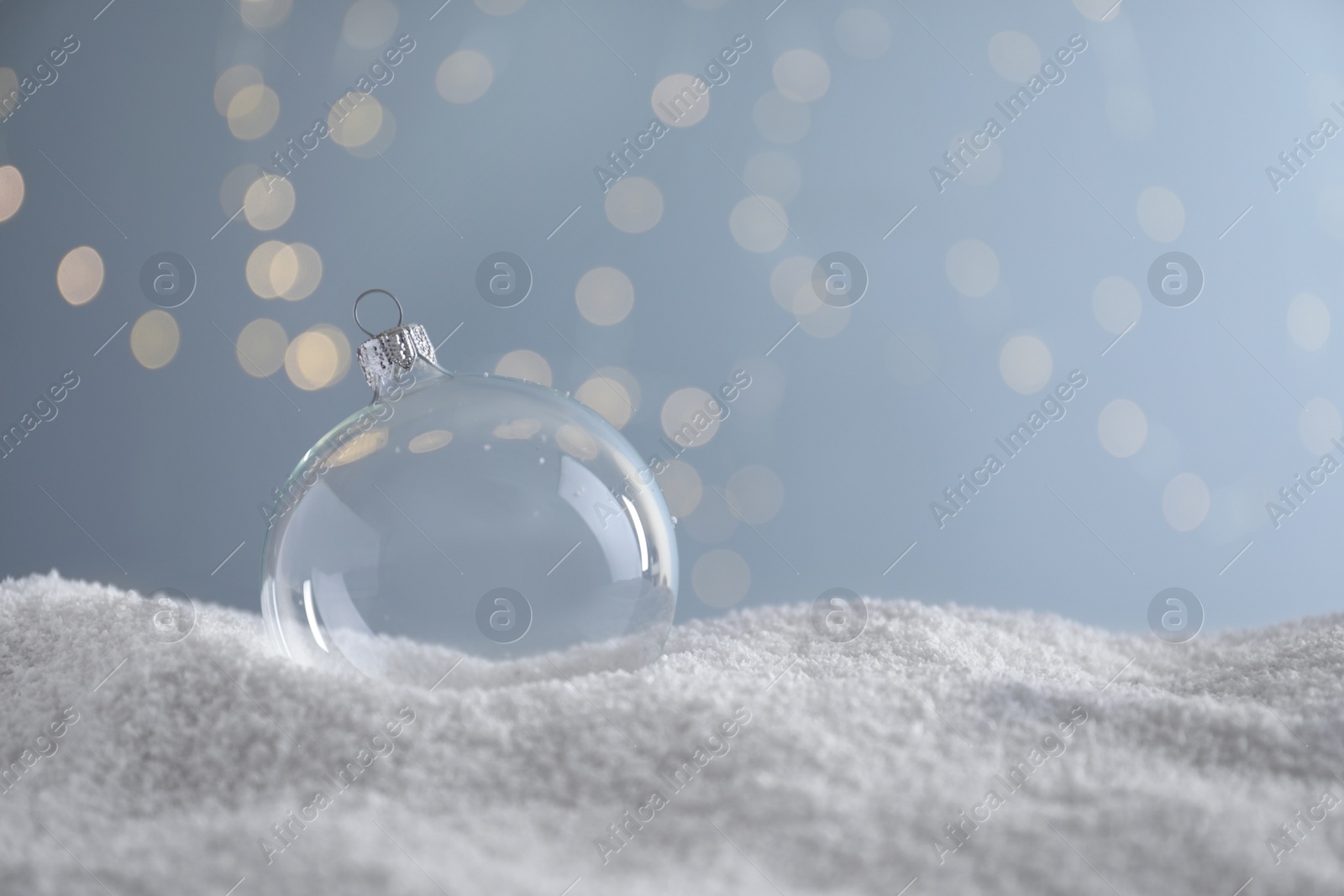 Photo of Transparent Christmas ball on snow against blurred fairy lights, space for text