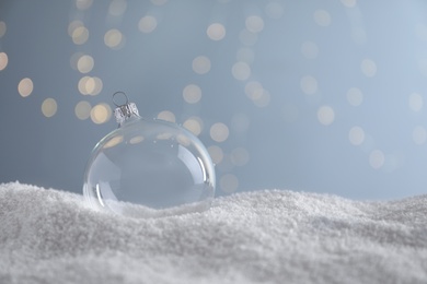 Photo of Transparent Christmas ball on snow against blurred fairy lights, space for text