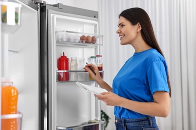 Young woman with notebook and pen near modern refrigerator indoors