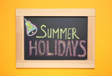 Photo of Chalkboard with phrase SUMMER HOLIDAYS on orange background, top view. School's out