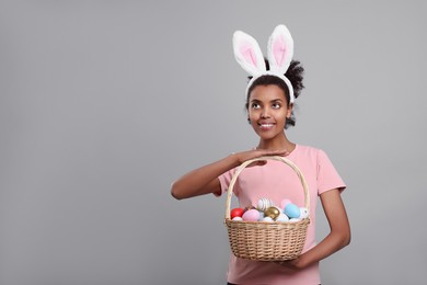 Happy African American woman in bunny ears headband holding wicker basket with Easter eggs on gray background, space for text