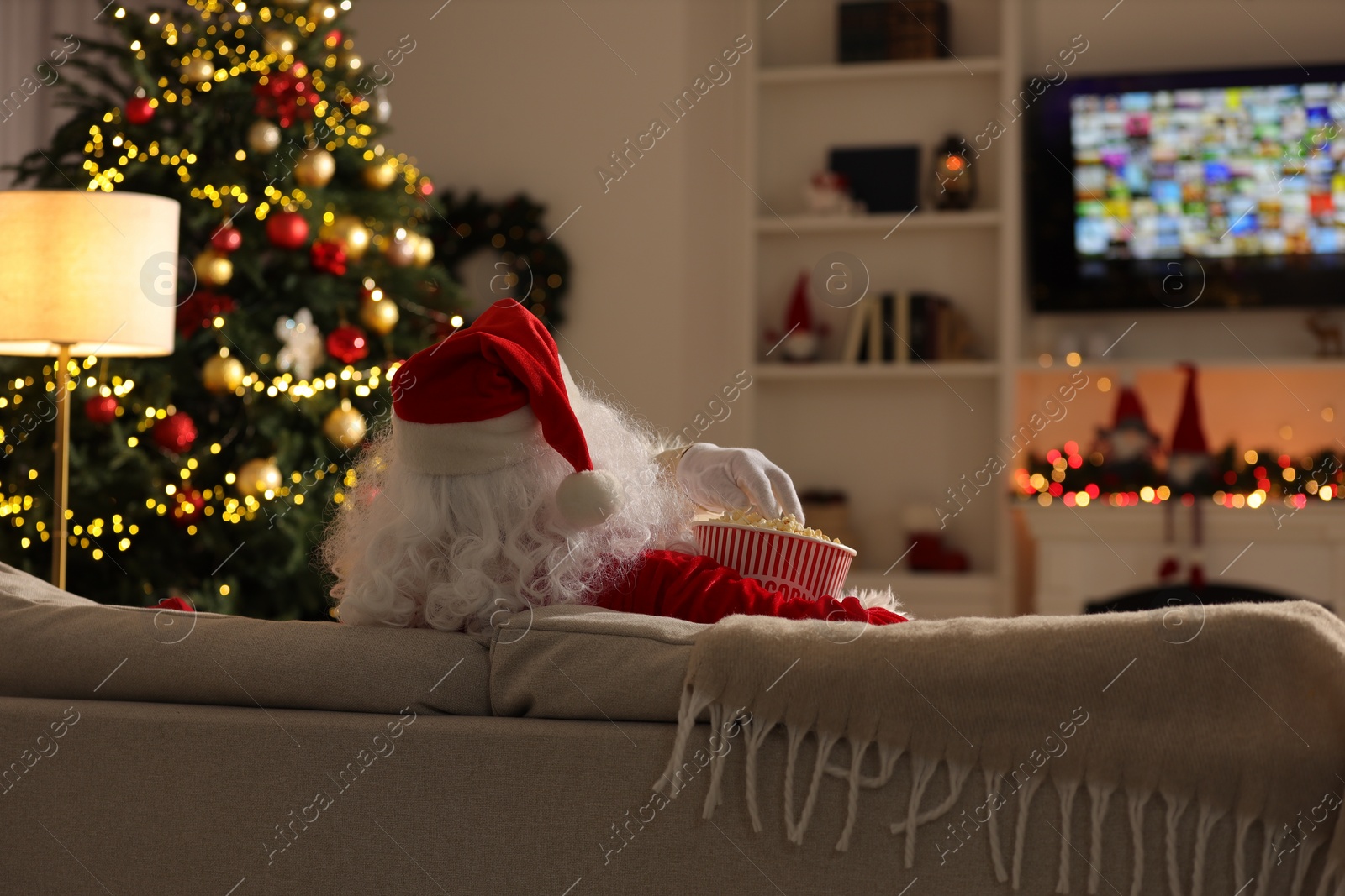 Photo of Merry Christmas. Santa Claus with popcorn bucket watching TV on sofa at home, back view