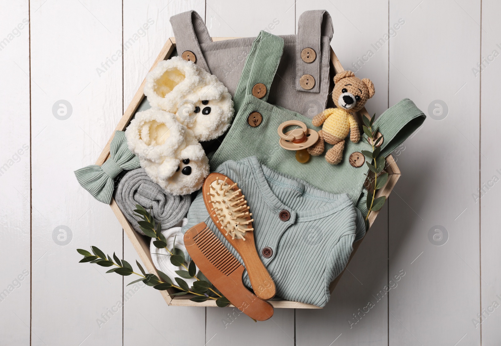 Photo of Children's clothes, shoes, toy bear and pacifier in crate on white wooden table, top view