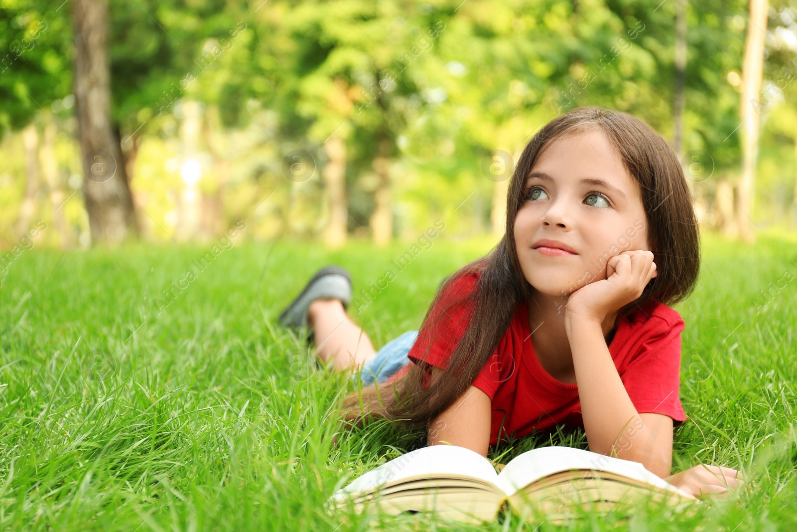 Photo of Cute little girl reading book on green grass in park