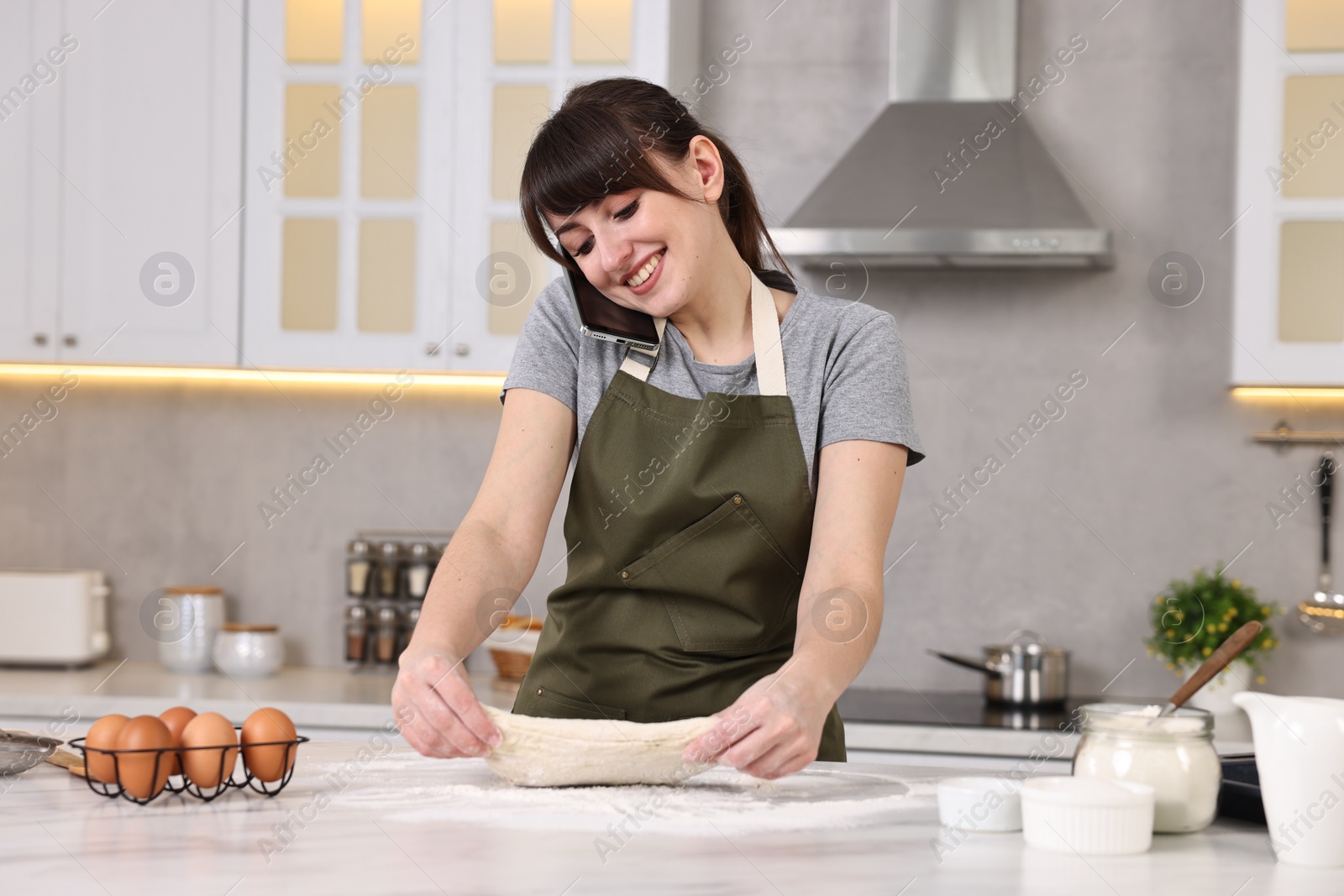 Photo of Happy young housewife kneading dough while talking on smartphone at white marble table in kitchen