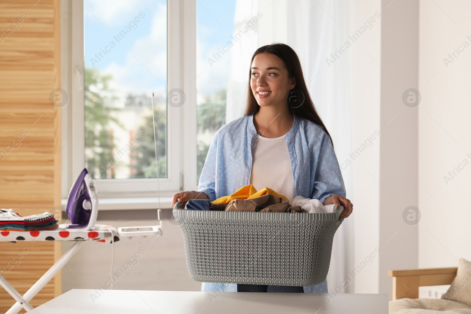 Photo of Young woman with basket full of clean laundry at table indoors