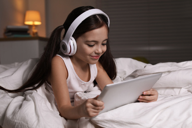 Photo of Cute little girl with headphones and tablet listening to audiobook in bed at home