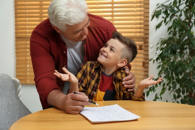 Photo of Little boy with his grandfather solving sudoku puzzle at table indoors