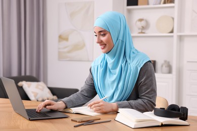 Muslim woman writing notes near laptop at wooden table in room