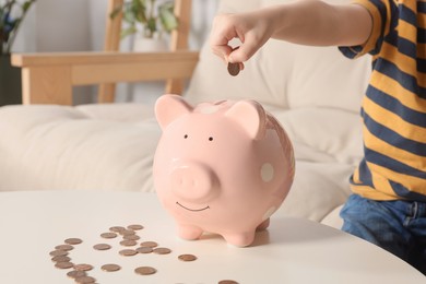 Little boy putting coin into piggy bank at table indoors, closeup